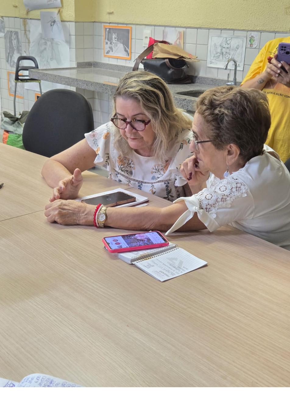 Duas artesãs olhando juntas para a tela de um celular, sobre a mesa de trabalho.