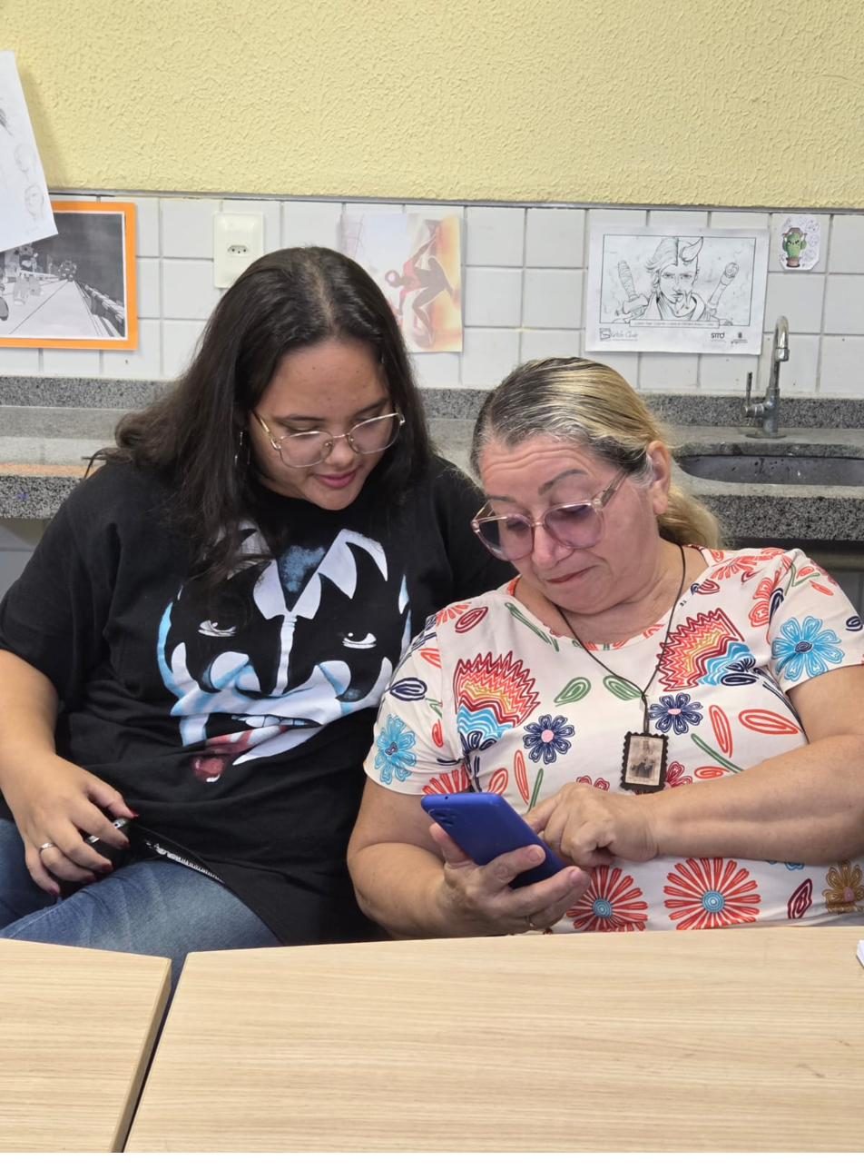 Voluntária mostra algo no celular para uma participante em sala de aula.