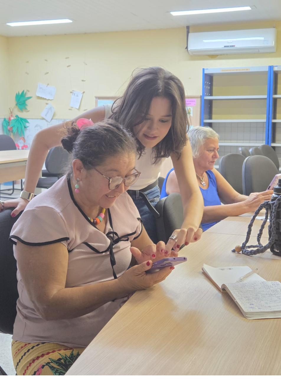 Voluntária ajuda uma participante a mexer no celular, com outra participante ao fundo.